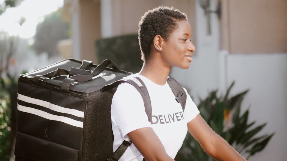 A young woman with short curly hair and a focused expression is outdoors, wearing a white t-shirt with the word 'DELIVERY' printed on it and carrying a black insulated delivery bag with white stripes 