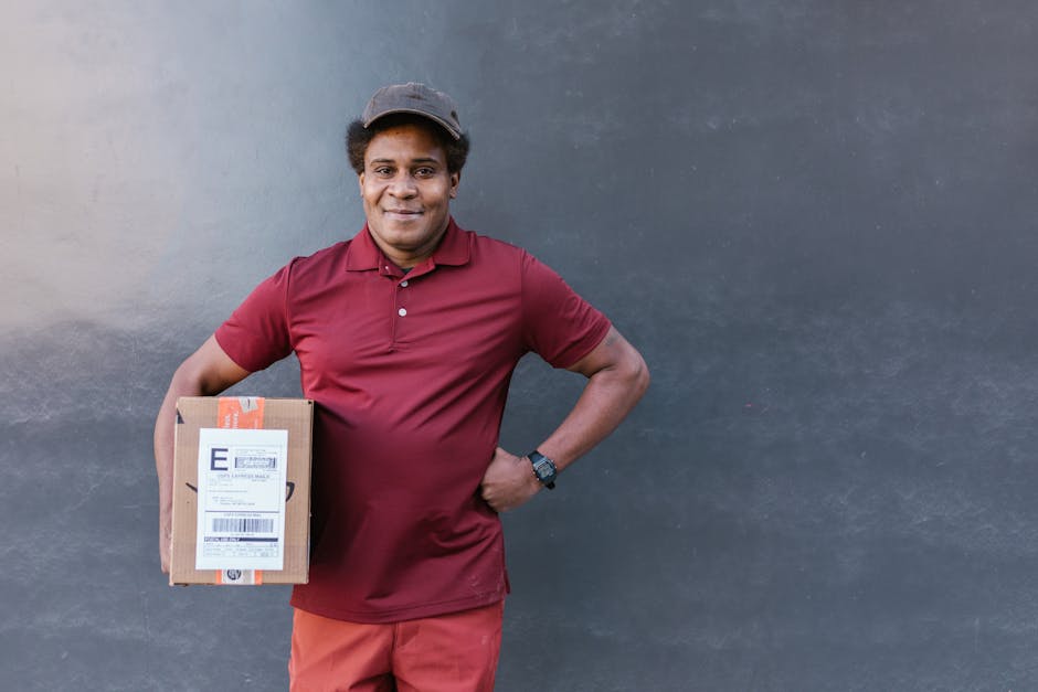 A male delivery driver with medium brown skin and short curly hair, wearing a dark gray cap, a maroon polo shirt, and matching pants, stands outdoors against a plain gray wall. He is smiling and holdi