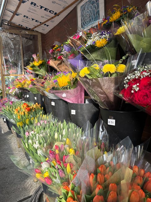 A display of various fresh flower bouquets arranged in black buckets at a flower shop, featuring a range of vibrant colors including yellow, purple, red, pink, orange, and white. The bouquets are wrap
