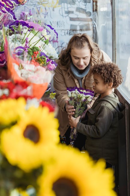 A woman and a young boy are inside a flower shop, standing near a large window illuminated by natural light. The woman, with light brown hair styled in loose waves, is wearing a beige coat and a light